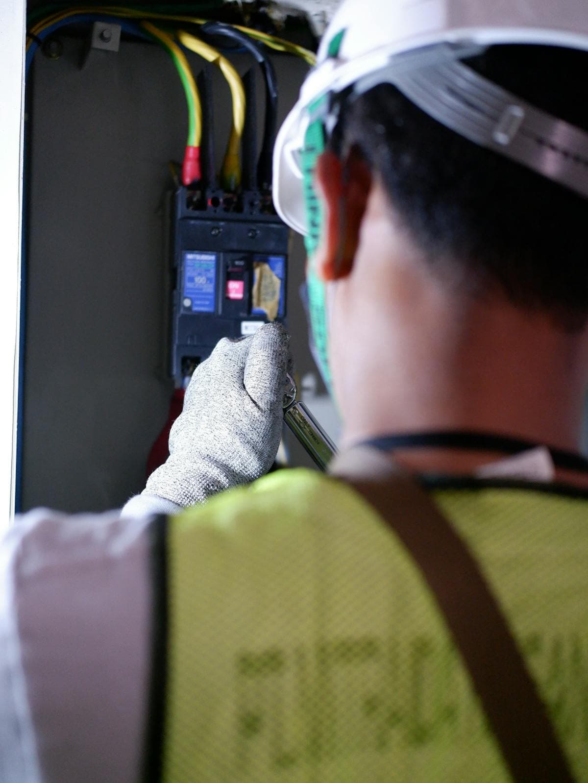 Electrician in a helmet, goggles, and hi-vis vest working on an electrical panel.